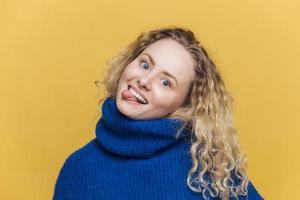 Funny comic curly young woman with positive expression, shows tongue, has fun with friends, wears blue sweater, makes grimace, isolated over yellow studio background. People and fun concept. photo