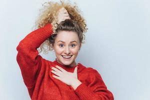 Adorable excited young female with curly hair, keeps hair tied in pony tail, dressed in casual red sweater, happy to recive positive news from interlocutor, poses against white studio background. photo
