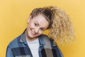 Funny cute female model with curly blonde pony tail, has positive smile, has fun alone indoor against yellow background, wears casual checkered shirt, being in good mood. Facial expressions. photo