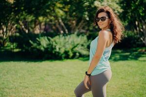 la mujer europea morena complacida en ropa activa posa de lado usa sunglases smartwatch tiene fitness al aire libre descansa después de trotar sonríe agradablemente. salud cuidado del cuerpo deporte y ejercicio concepto foto