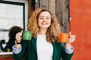 Portrait of cute female with pure skin having stylish hairstyle dressed in green jacket and white T-shirt holding two cups of tea going to give one to her boyfriend having pleased, excited expression photo