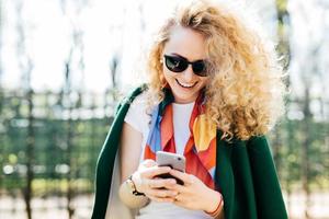 hermosa joven en tonos redondos de moda enviando mensajes de texto a sus amigos a través de las redes sociales mientras está de pie afuera en un parque verde disfrutando del buen tiempo soleado foto