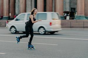 Outdoor shot of happy smiling woman spends time in active way rides on skating rollers demonstrated high speed during motion poses on roaad with transport in background. Hobby and lifestyle. photo