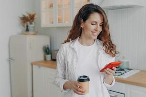European woman with smartphone has coffee. Girl is reading message in kitchen at home and smiling. photo