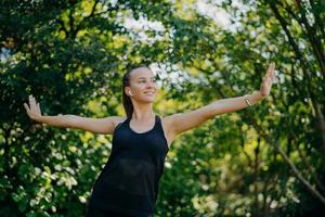 Training outdoors concept. Pleased sporty European woman feels freedom motivates you for sport stretches arms sideways leads active lifestyle wears black t shirt poses against green trees outside. photo