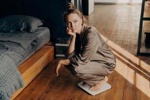 Young upset woman in satin pajama measuring her weight at home photo