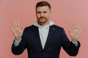 Serious confident adult man with bristle raises hands towards camera shows palms has strict expression asks to hold on or stop wears comfortable hoodie and jacket isolated over pink background photo