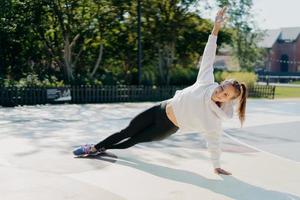 Glad energetic dark haired woman stands in sideways plank keeps arm raised strengthens core muscle improves stability has workout outdoor wears sportsclothes enjoys sunny day. Endurance concept photo