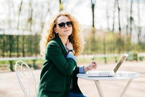 Caucasian female freelancer with fluffy blonde hair wearing trendy clothes and sunglasses working on modern laptop pc holding hand on her neck being little tired and exhausted isolated over nature photo