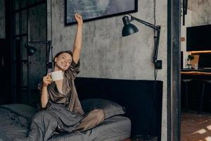 Young beautiful sleepy woman with hair tied up in bun starting her morning with cup of hot coffee photo