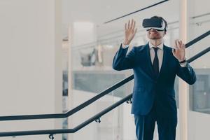 Impressed male office worker wearing VR goggles exploring virtual world while standing in office photo