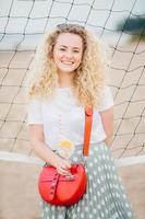 Vertical shot of good looking curly female wears t shirt and skirt, has small bag, holds cocktail with ice, stands near tennis net on beach, has good rest. Lovely woman with appealing charming smile photo