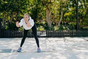 una foto al aire libre de una mujer deportiva sana y satisfecha se inclina hacia adelante mantiene las palmas levantadas hace ejercicios en trenes al aire libre en el parque se siente bien usa sudaderas con mallas y zapatillas lleva un estilo de vida activo