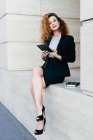 Vertical portrait of pretty slim woman with curly hair, wearing black jacket, skirt and high-heeled shoes, using modern tablet computer for searching information for her work, writing in notebook photo