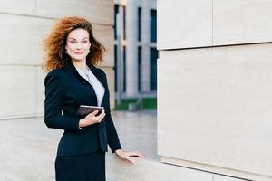 Pleasant-looking female dressed formally, holding digital tablet computer which is necessary for her work. Beatiful woman with curly bushy hair, having attractive appearance, going for her work photo