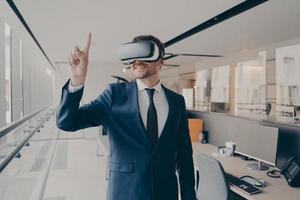 Happy man office worker wearing vr goggles, pointing with finger at objects in digital world photo