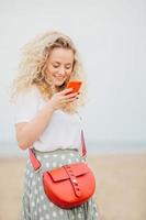 Satisfied young female with bushy curly hair holds smart phone, uses free internet connection for texting messages, stands against blurred seascape background. People and recreation concept. photo