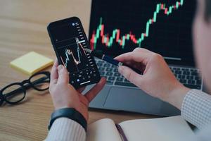 Man is pointing with pen to financial chart phone screen. PC on the desk at workplace in office. photo