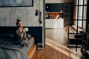 Young happy attractive girl sitting on bed in lotus position and drinking morning coffee photo