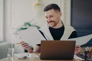 Handsome happy businessman analyzing documents and feeling satisfied while working on laptop from home photo