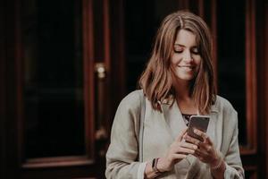 Horizontal shot of pretty cute brunette female model shares multimedia files via cell phone, dressed in elegant clothes, reads positive news in internet website, connected to wireless internet. photo