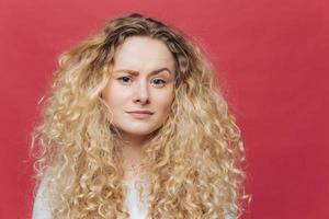 Young attractive woman has discontent expression, raises eyebrow in bewilderment, being dissatisfied with something, has bushy curly hair, isolated over pink background. Facial expressions concept photo