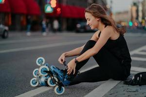 tiro al aire libre de mujer activa cordones patines se prepara para el paseo se sienta en la carretera contra el fondo de la ciudad ocupada vestida con ropa deportiva negra disfruta patinando. estilo de vida deportivo y concepto de recreación foto