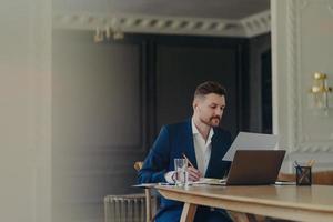 Serious businessman in smart suit writes in notepad concentrated at paper document poses at office desk with laptop computer glass of water uses wireless earphones for listening information. photo