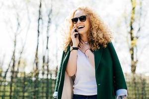 retrato de medio perfil de mujer atractiva con cabello rizado usando ropa elegante y gafas de sol hablando por teléfono inteligente, sonriendo y riendo con expresión feliz y emocionada tocando su cuello foto