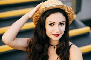 Beautiful woman with dark eyes and hair having nice make-up wearing straw hat sitting at stairs having confident look. Attractive brunette female with appealing appearance posing into camera photo