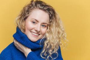 Close up shot of delighted satisfied female model has curly blonde hair, broad smile, demonstrates white perfect teeth, wears blue knitted sweater, isolated over yellow background. Happiness photo