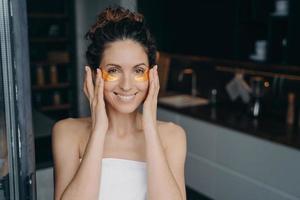 una joven bonita envuelta en una toalla después de bañarse es feliz y relajante aplicando parches en los ojos. foto