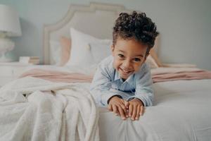 Happy curly afro american boy laying on bed and smiling at camera photo