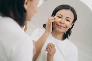 Smiling female apply under eye patches, looking at mirror. Skincare treatment routine, self-care photo