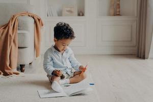 Small curly afro mulatto boy with colouring book at home photo