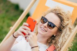 Young curly blonde woman lies in hammock, enjoys summer day, bloggs and chats via smart phone, wears trendy shades, has positive smile, recreats during holidays. People and lifestyle concept. photo