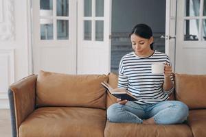 Woman rests reading bestseller novel holding paper book, coffee cup, sitting on sofa at home photo