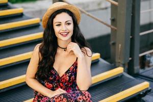 Smiling female with dark luxurious hair wearing dress and straw hat sitting at stairs looking with delightful look into distance. Restful young woman with appealing appearance sitting outdoors photo