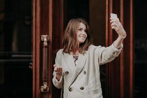 Adorable female student poses for making selfie, uses modern cell phone, dressed in white jacket, stands near entrance to building, has happy expression. People, modern technologies concept. photo