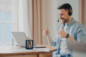 Satisfied bearded male model attends online training from home uses headset makes like gesture with both hands looks attentively at laptop computer makes video call poses against cozy interior photo