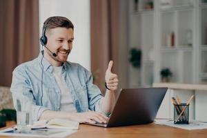 I like it. Glad bearded adult man has video conference keeps thumb up likes something focused at laptop computer has distance job interview dressed casually. Professional call center agent working photo