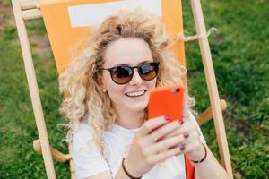 Positive European female with friendly smile, uses modern electronic device, happy to recieve message from boyfriend, rests outdoor during summer vacation. People, recreation, lifestyle concept photo