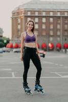 Vertical shot of slim woman wears cropped top and trousers poses on rollers enjoys free time has healthy body enjoys rollerblading poses outdoor against urban background. Lifestyle activity.