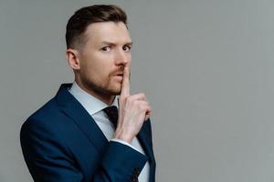 Serious bearded adult man makes shush gesture asks to be quiet tells confidential information presses index finger to lips wears formal clothes isolated over grey background with copy space. photo