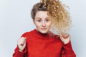 Horizontal shot of beautiful blue eyed funny female with curly bushy hair tied in pony tail, keeps hands pressed in fists, raises them, has unexpected reaction on something shocking, isolated on white photo