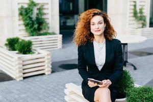 Beautiful gorgeous woman with wavy bushy hair, wearing formal suit, sitting crossed legs at outdoor cafe, holding tablet computer while waiting for someone, looking at camera with confident look photo