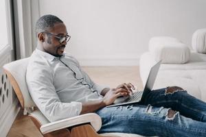 Online freelance job. Handsome african american man in cosy chair in front of pc at his apartment. photo
