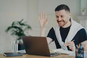 Cheerful bearded man has video conference online waves palm in laptop computer uses wireless earphones poses at coworking space dressed casually writes down information in diary smiles gladfully photo