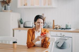 Happy girl is sitting at the table at kitchen and texting on phone. Workplace at home. photo