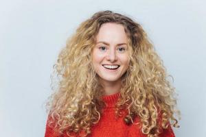 una foto aislada de una atractiva y alegre mujer rubia y hermosa tiene el pelo rizado, tiene una amplia sonrisa, está de buen humor después de un día exitoso, posa contra un fondo de estudio blanco en blanco. mujer joven encantada